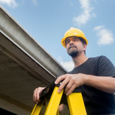 goateed roofer in black shirt and hardhat standing on a yellow ladder